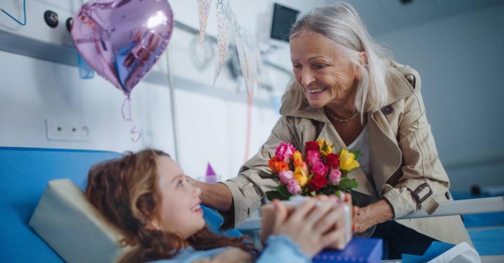 An elderly woman visiting her granddaughter at the hospital. She is giving her granddaughter flowers and a present.