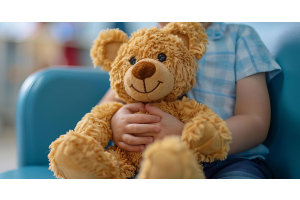 A child is holding a stuffed teddy bear. They are patiently in a waiting office and sitting in a large blue chair.