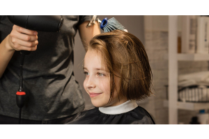 A little girl in a hair salon chair getting her hair cut. The hairstylist is blow drying the girl’s freshly cut bob haircut.