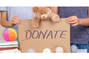 Two hands holding up a cardboard sign that reads “Donate.” A brown stuffed teddy bear leans over the top of the sign.