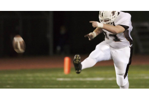 A high school football player kicking a football. He is playing the game at night on an illuminated football field.