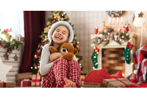 A little girl hugging a teddy bear. She sits on the floor in her living room, which is full of holiday decorations.