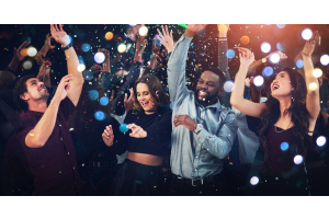 Four adults dancing in a dark setting. There are bright lights illuminating the group and confetti falling from the roof.