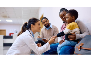 A pediatrician talking to a little boy sitting on his mother’s lap. The pediatrician is kneeling and holding his hand.