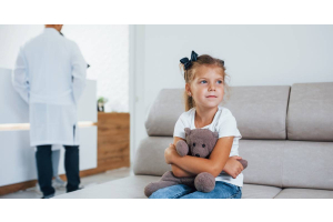 A little girl sitting on a chair, holding a teddy bear. A man in a white suit stands behind her, talking to a receptionist.