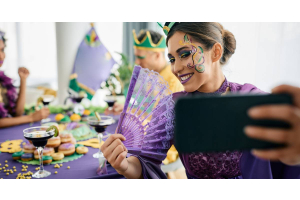 A woman taking a photo of herself while sitting at a Mardi Gras party. She is wearing a purple gown and holding a fan.