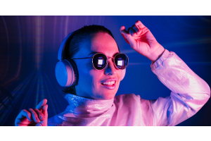 A smiling woman wearing round sunglasses and silver headphones dancing while illuminated by neon pink and blue lights.