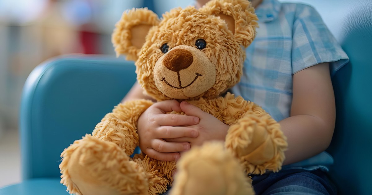 A child is holding a stuffed teddy bear. They are patiently in a waiting office and sitting in a large blue chair.