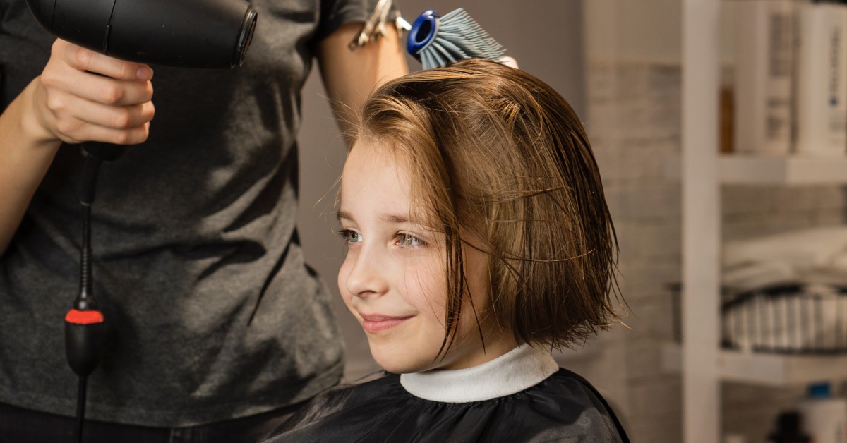 A little girl in a hair salon chair getting her hair cut. The hairstylist is blow drying the girl’s freshly cut bob haircut.