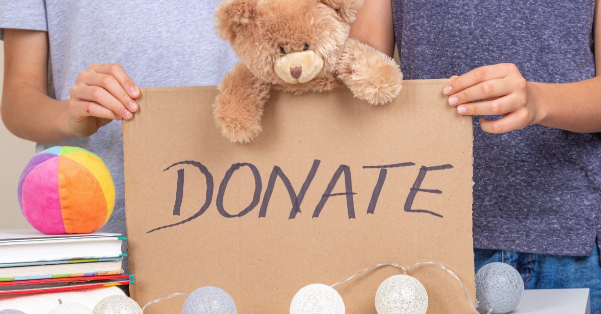 Two hands holding up a cardboard sign that reads “Donate.” A brown stuffed teddy bear leans over the top of the sign.