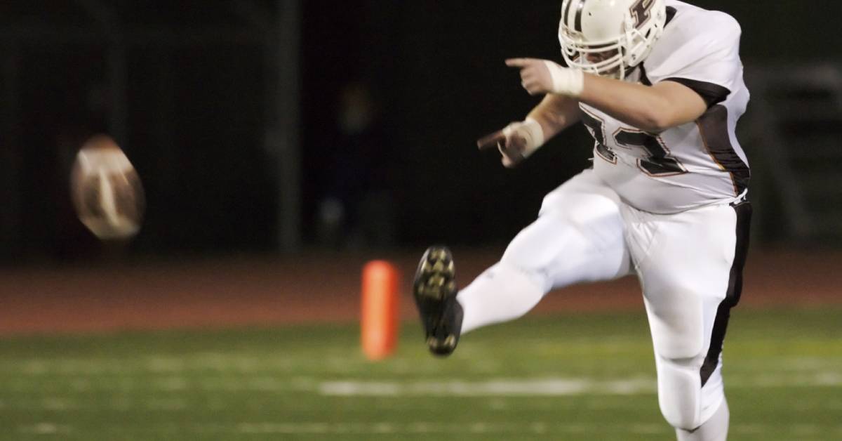 A high school football player kicking a football. He is playing the game at night on an illuminated football field.