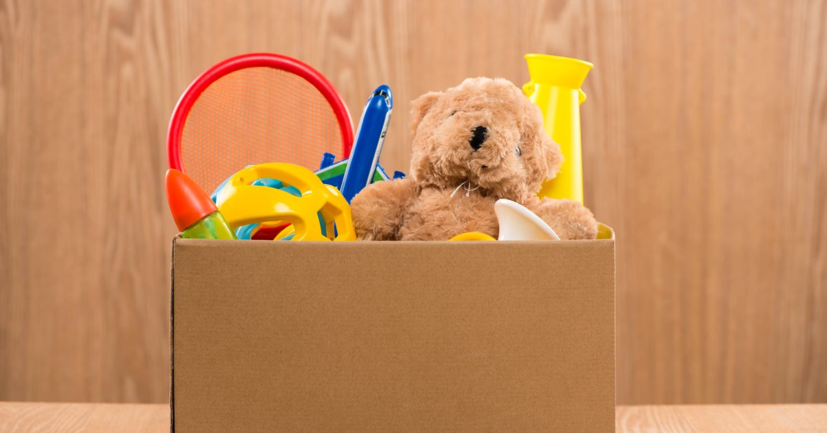 A cardboard box filled with children’s toys. There are plastic toys and a teddy bear sticking out from the top.