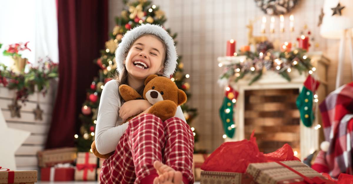 A little girl hugging a teddy bear. She sits on the floor in her living room, which is full of holiday decorations.