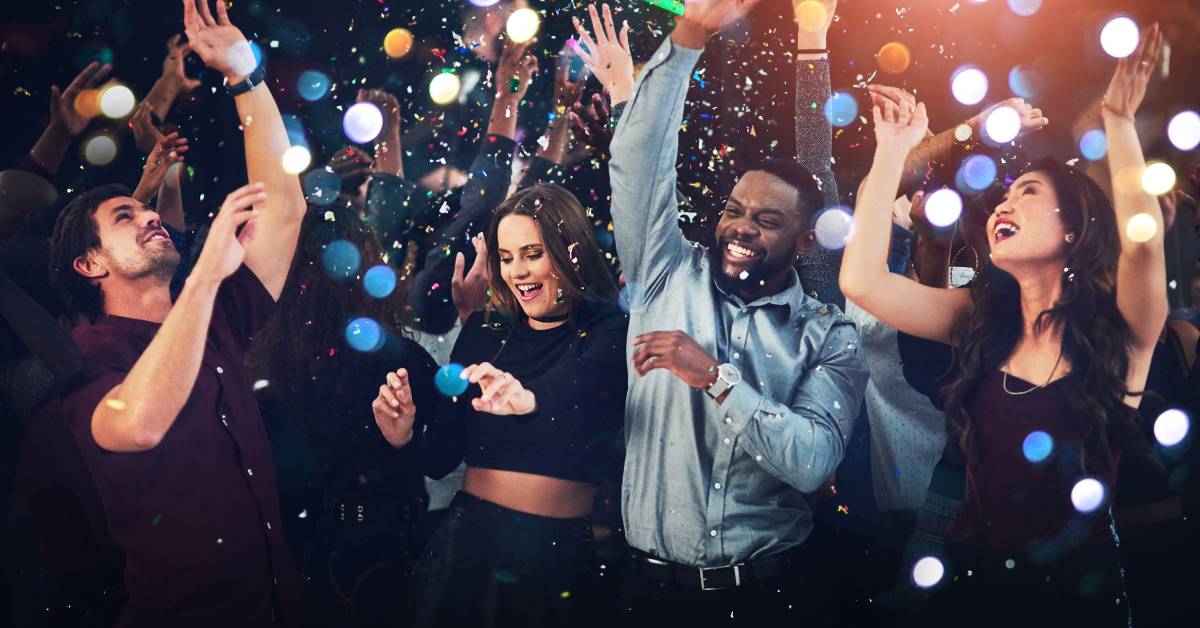 Four adults dancing in a dark setting. There are bright lights illuminating the group and confetti falling from the roof.