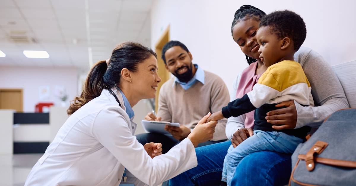 A pediatrician talking to a little boy sitting on his mother’s lap. The pediatrician is kneeling and holding his hand.