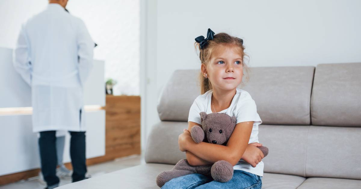 A little girl sitting on a chair, holding a teddy bear. A man in a white suit stands behind her, talking to a receptionist.