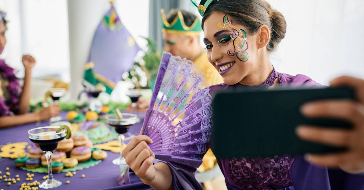 A woman taking a photo of herself while sitting at a Mardi Gras party. She is wearing a purple gown and holding a fan.