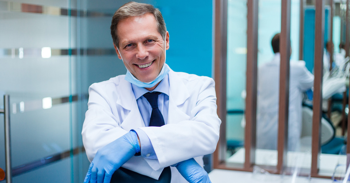 middle-aged male dentist smiling in his dentist's office, wearing a white lab coat, blue button down, black tie, and blue latex gloves