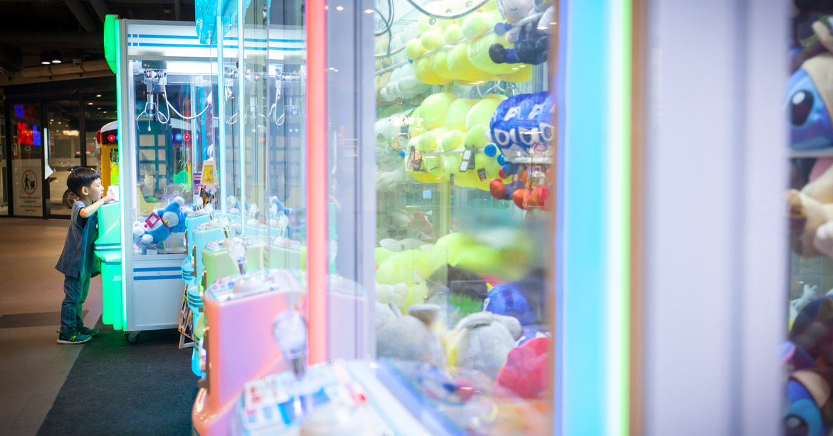 A close-up of an arcade game full of plush toys, with a young boy playing an arcade claw game in the background.