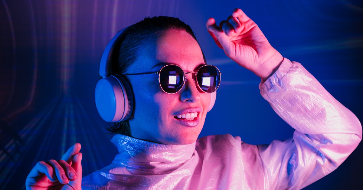 A smiling woman wearing round sunglasses and silver headphones dancing while illuminated by neon pink and blue lights.