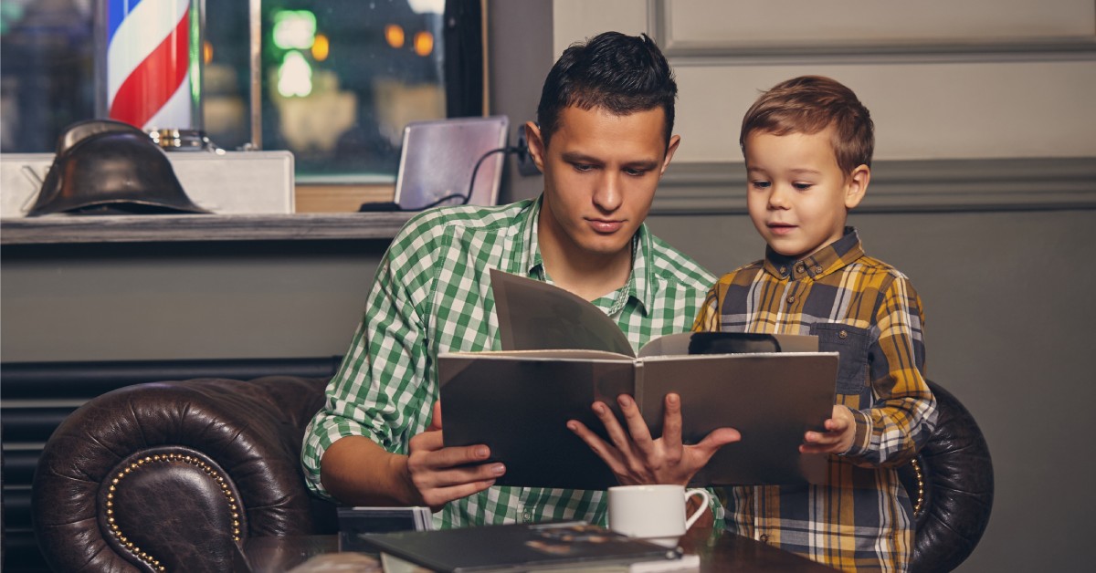 A child and his dad reading from a book in a barbershop waiting area. The dad is showing his son pictures in the book.