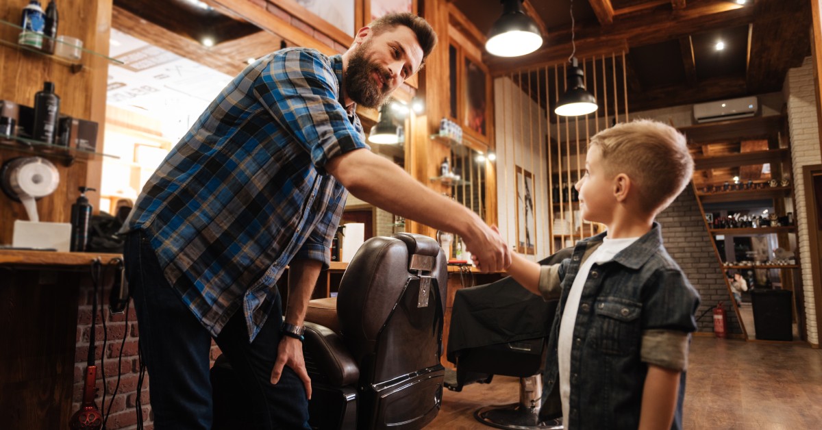 A hairstylist shaking hands with his young client at a hair salon. The young client is happy with his new buzzed haircut.