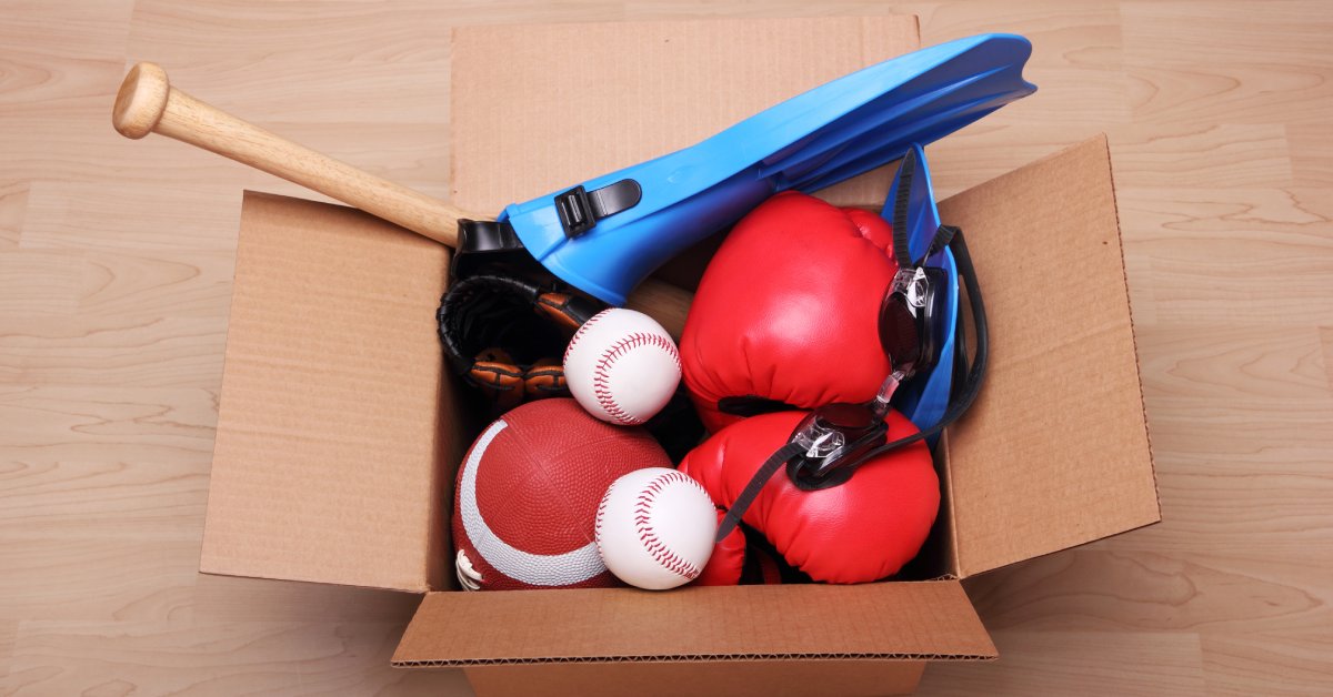 A box overflowing with various sports equipment, such as a baseball bat, baseballs, a football, boxing gloves.