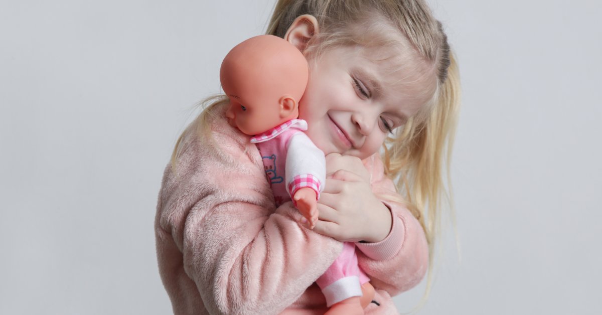 A little girl with blonde hair hugs a baby doll in pink and white clothes. The girl is smiling and closing her eyes.