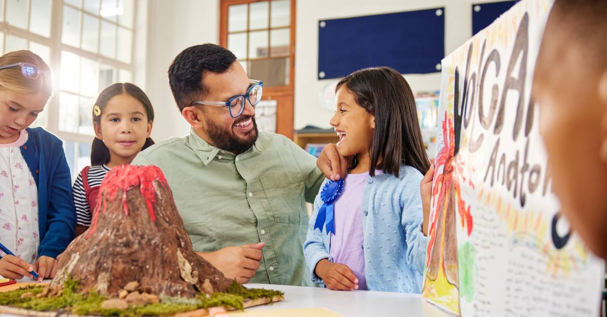 A teacher pinning a blue ribbon on his student’s clothes. The student is presenting a science project with a volcano.