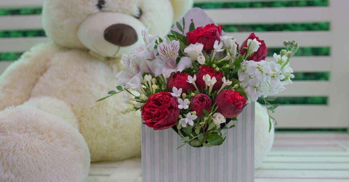 A floral arrangement inside of a square box. Behind the box is a large, beige teddy bear slumped over.