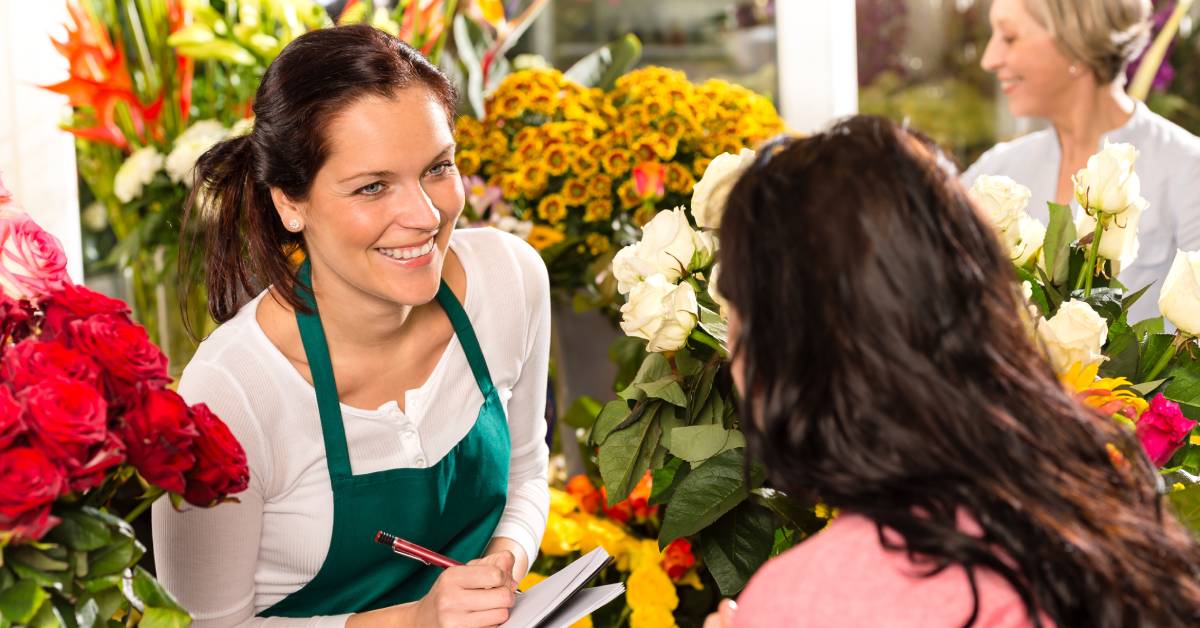 A florist holds a clipboard and a pen while talking to a customer. There are dozens of flowers on display surrounding them.