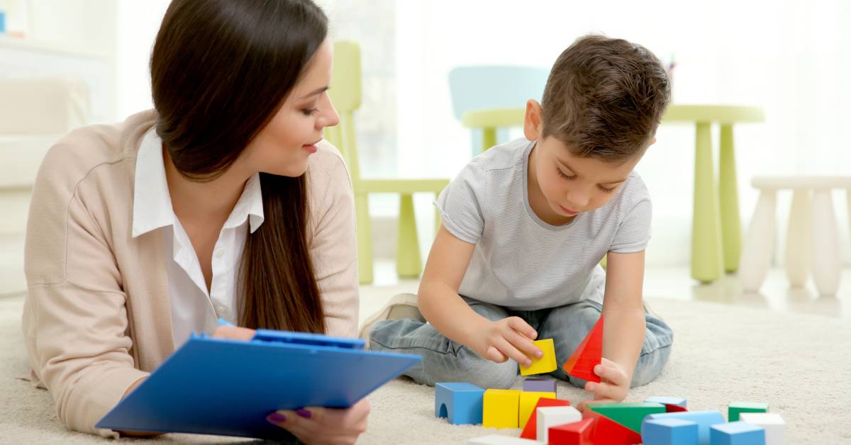 A little boy playing with building blocks on the floor. A woman is laying next to him with a clipboard in her hand.