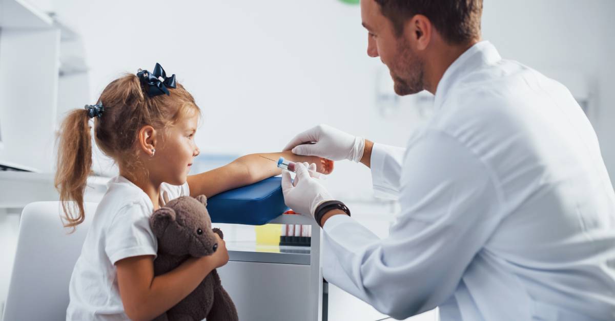 A doctor drawing a little girl's blood. The little girl holds a teddy bear in one arm, stretching the other out.