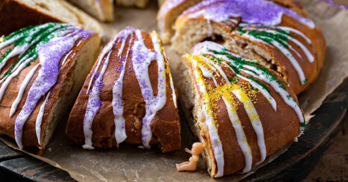 A King cake decorated in Mardi Gras colors. There is a small plastic baby toy sitting between the slices of cake.