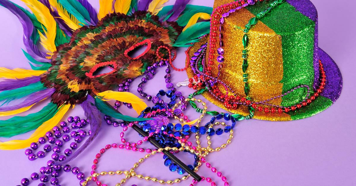 Mardi Gras party favors sitting against a purple background. There is a mask, a hat, and colorful beaded necklaces.